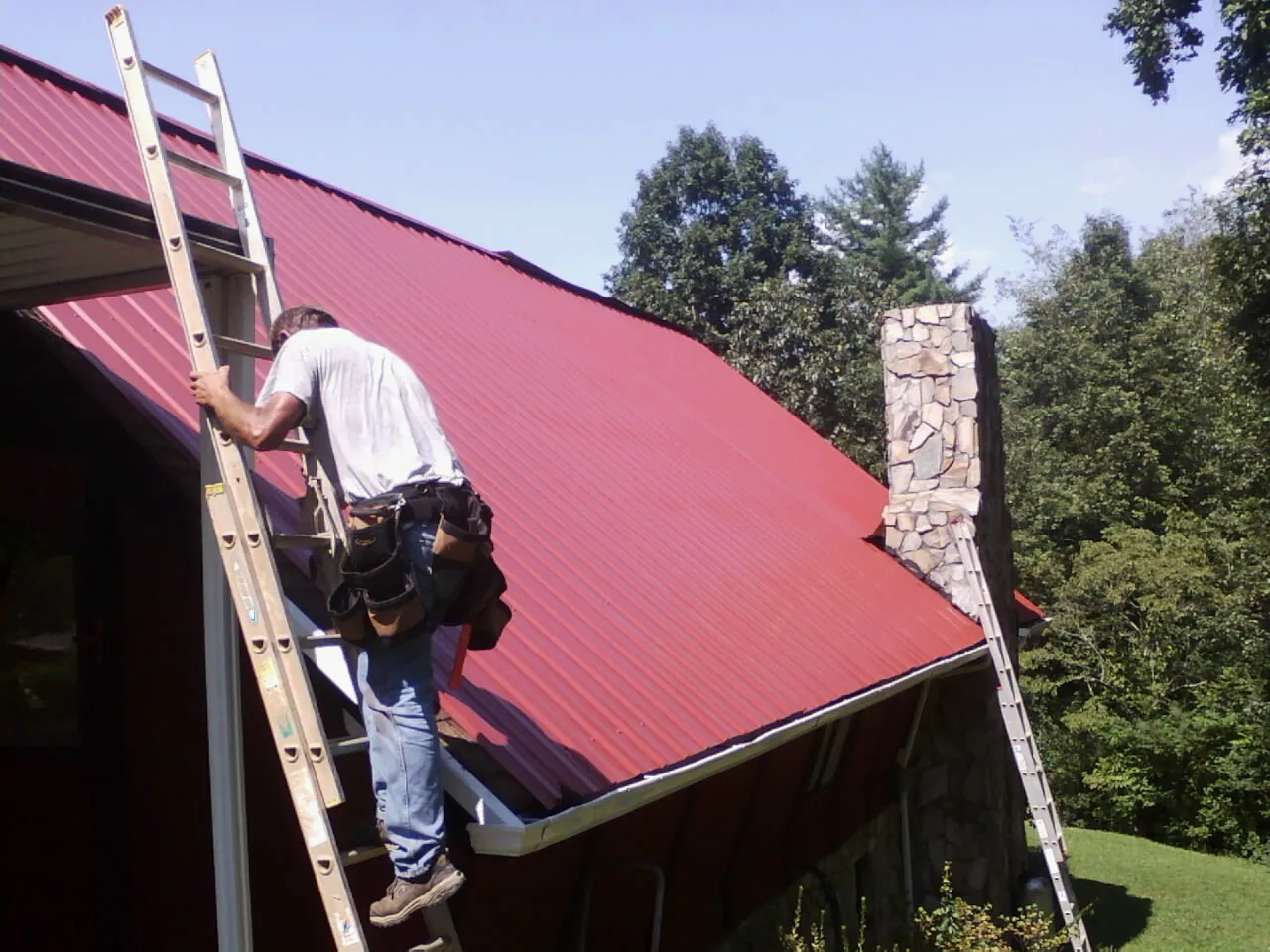 Roofer on a ladder beside a red metal roof and stone chimney — Soffit & Fascia Repair by Roofing Brothers in Valentines
